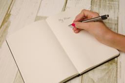 Thumbnail sized: Hand with pen over a journal on desk
