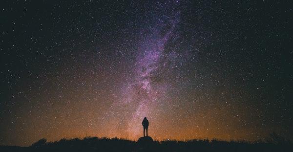 Small figure of a man against dark sky full of stars with milky way going down the center.